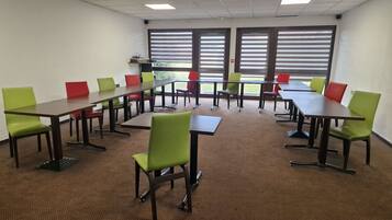 Un salón de conferencias con varias mesas y sillas, piso alfombrado y amplias ventanas con persianas.