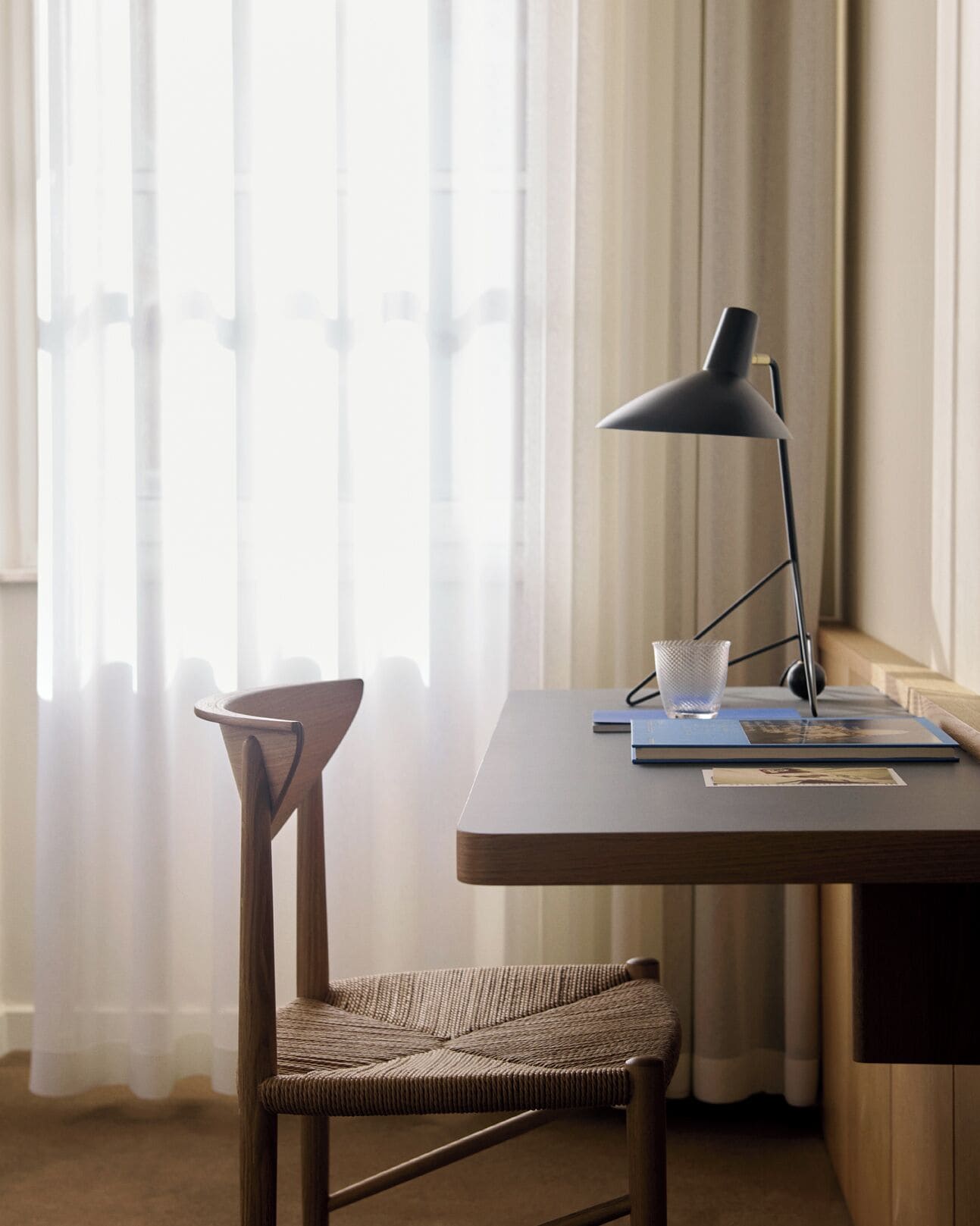 Een moderne kamer met een houten stoel, een bureau met een lamp en uitzicht door dunne gordijnen.
