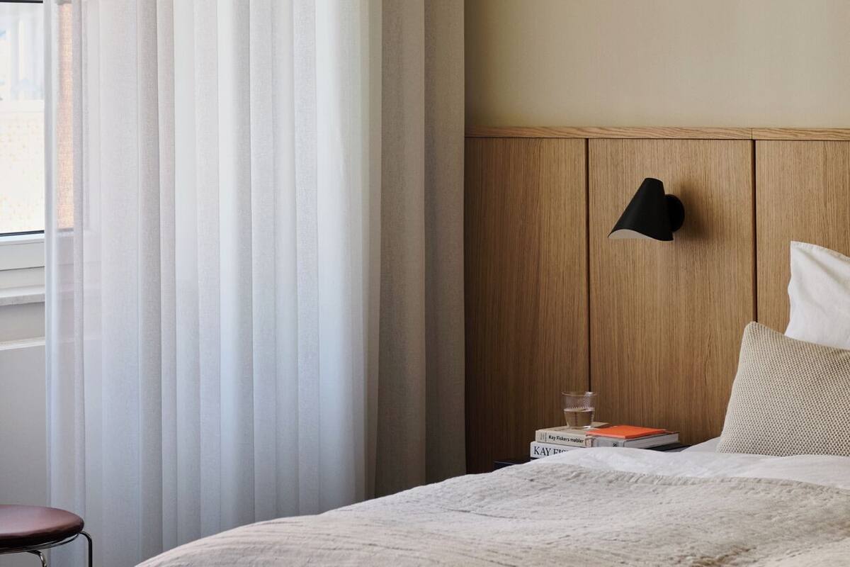 A hotel room with a bed, a bedside table, a lamp, and a window with curtains.