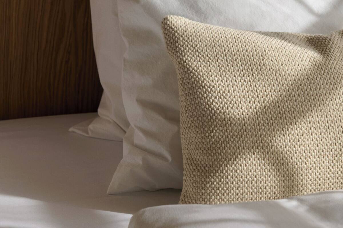 A bed with white linens and a textured beige pillow against a wooden headboard.