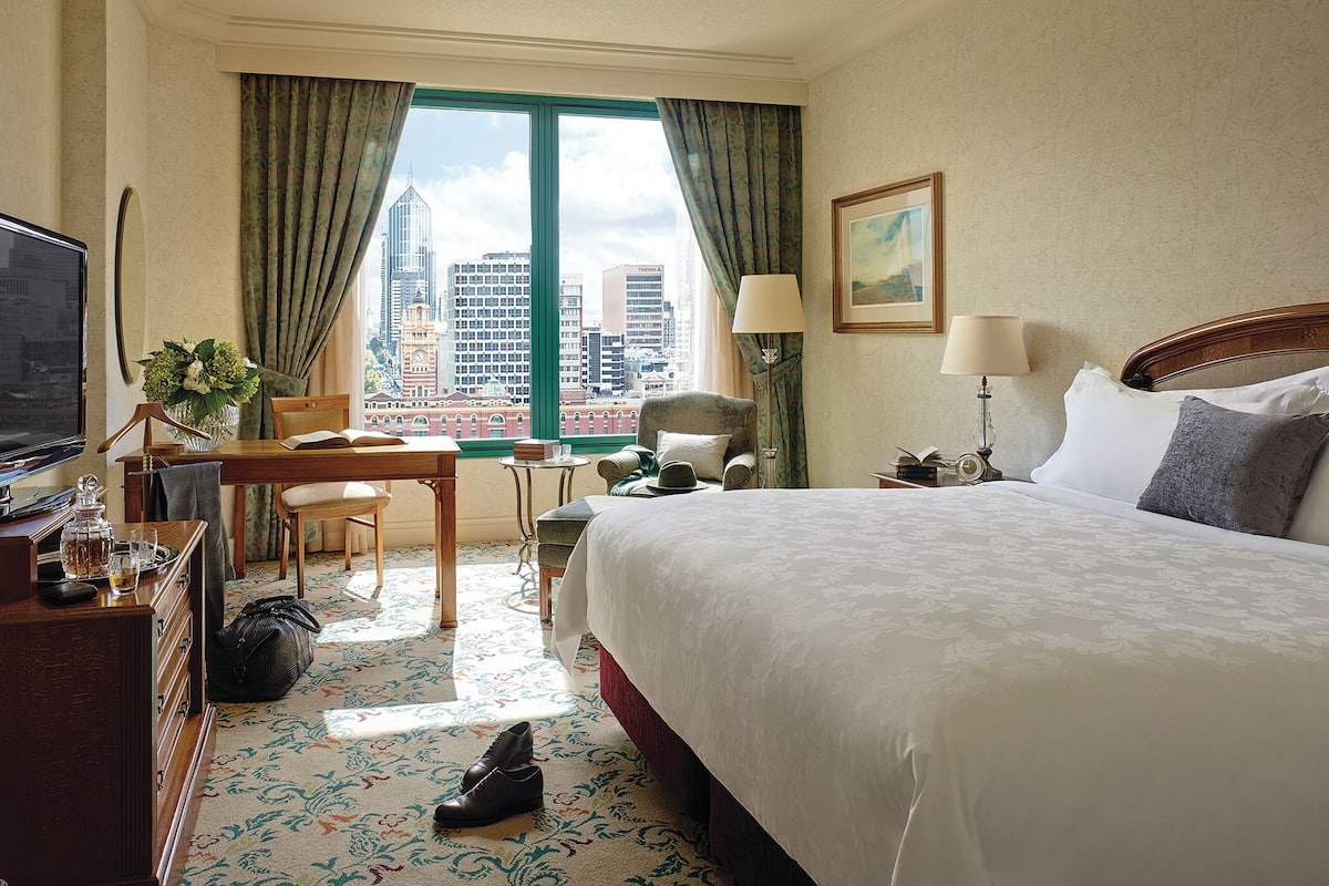 A hotel room with a large bed, a desk with a chair, a television, and a view of the cityscape through the window.