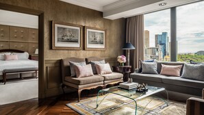 A modern hotel room with a large bed, a sofa, and a couch. There are two framed pictures on the wall, a lamp, and a view of the city through large windows.