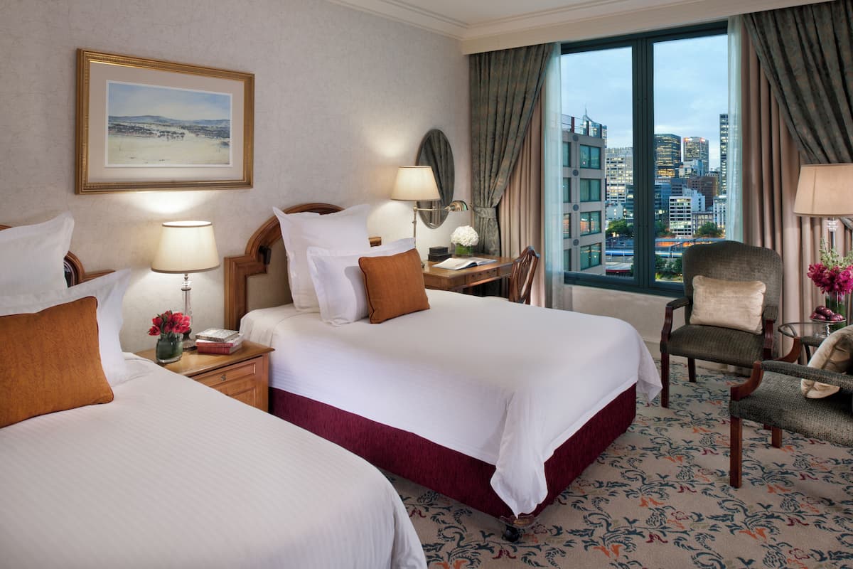 A hotel room with two beds, a desk, a chair, and a large window with a city view.