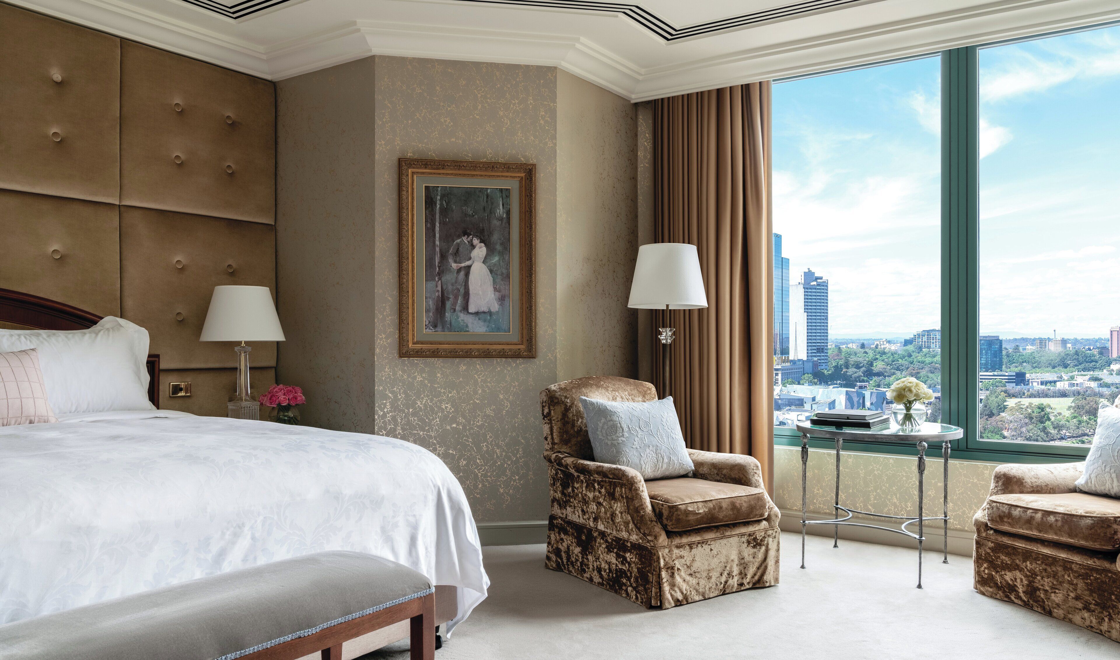Habitación de hotel con una cama grande, dos sillones, un cuadro en la pared y vistas de la ciudad a través de amplios ventanales.
