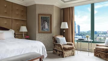 A hotel room with a large bed, two armchairs, a painting on the wall, and a view of the city through large windows.