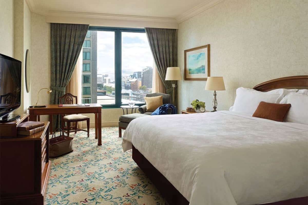 A hotel room with a large bed, a desk with a chair, a television, and a view of the city through large windows.