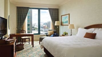 Habitación de hotel con una cama grande, un escritorio con silla, televisión y vistas a la ciudad a través de amplios ventanales.