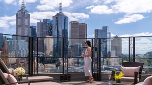 A rooftop terrace with a city view, furnished with wicker chairs and a table, and a glass barrier.