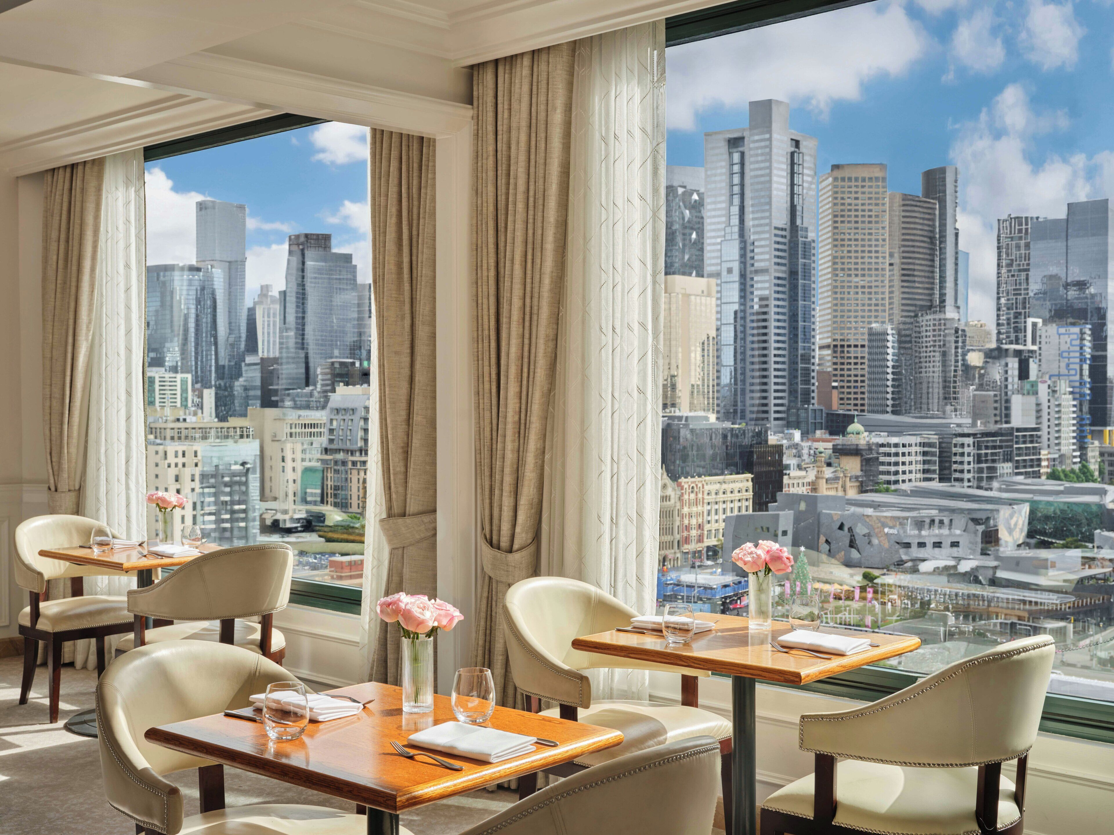 A hotel room with a city view, a dining area with tables and chairs, and a vase with flowers.
