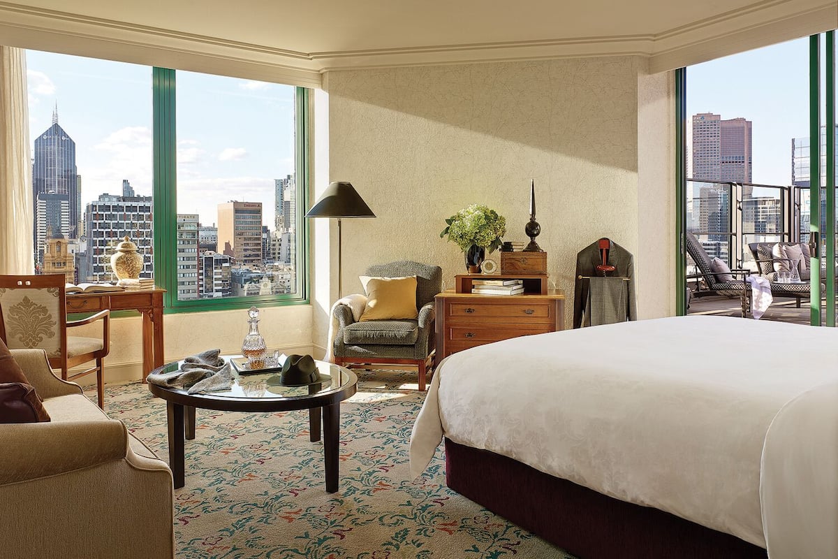 A hotel room with a large bed, a desk, a chair, and a view of the cityscape through large windows.
