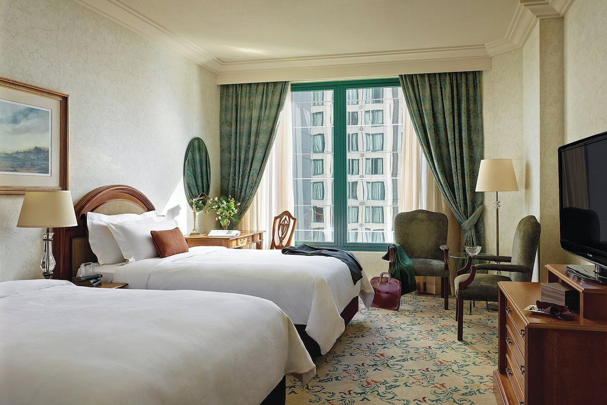 A hotel room with two beds, a television, a desk with a lamp, a chair, and a view of buildings through the window.