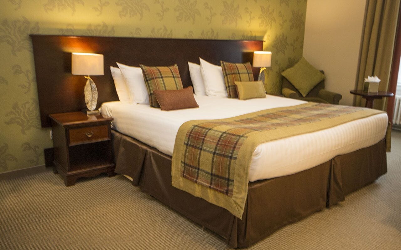 A hotel room with a large bed, two bedside tables with lamps, a nightstand, and a green patterned wall.