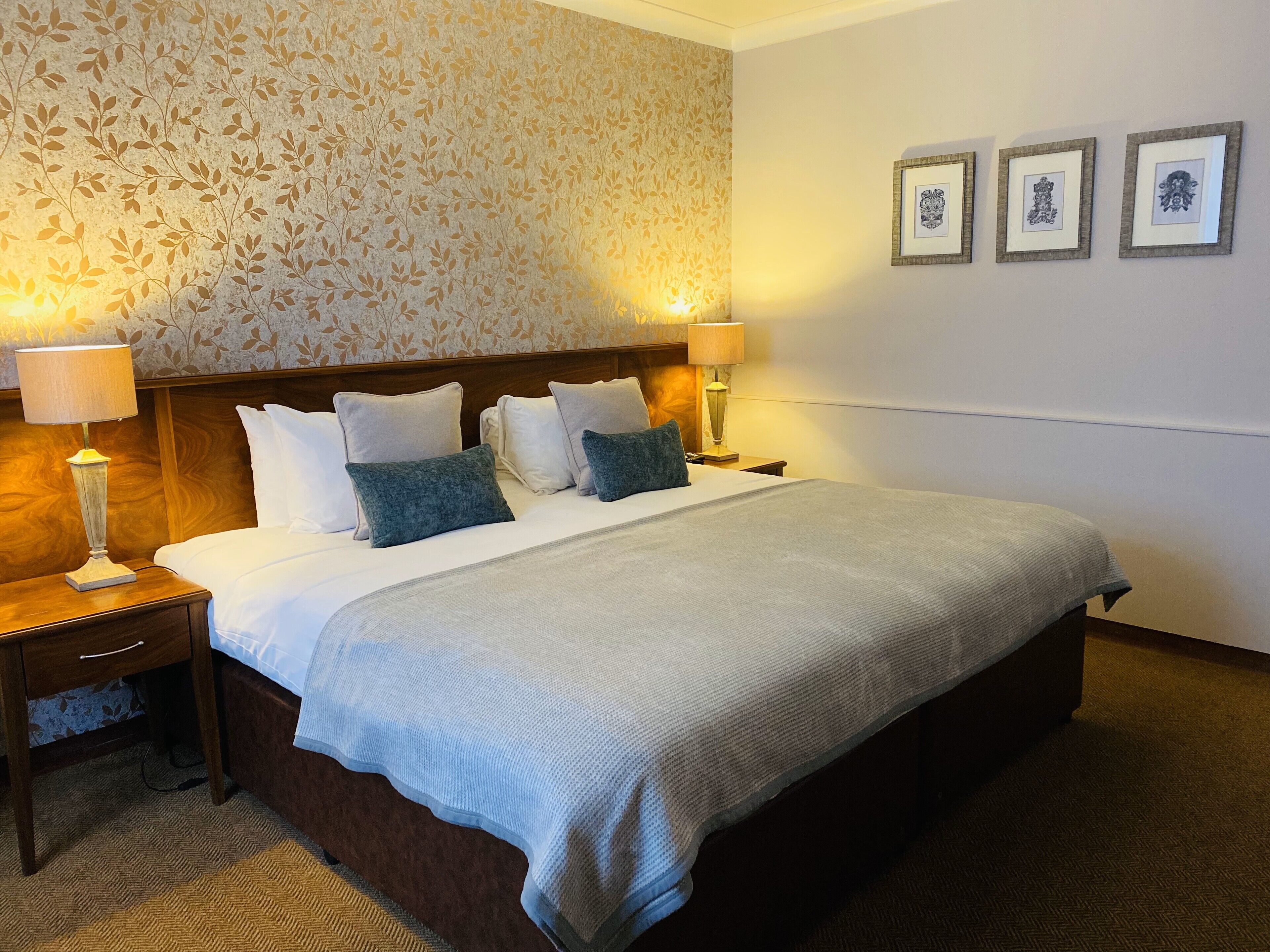 A hotel room with a large bed, two bedside tables, a nightstand, a patterned wallpaper, and three framed pictures on the wall.
