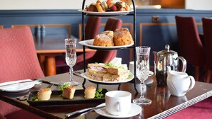 Un plateau à trois niveaux, garni de sandwichs, de scones et d’une tasse de thé, est posé sur une table dans un coin repas, où l’on trouve des chaises rouges et un mur bleu.