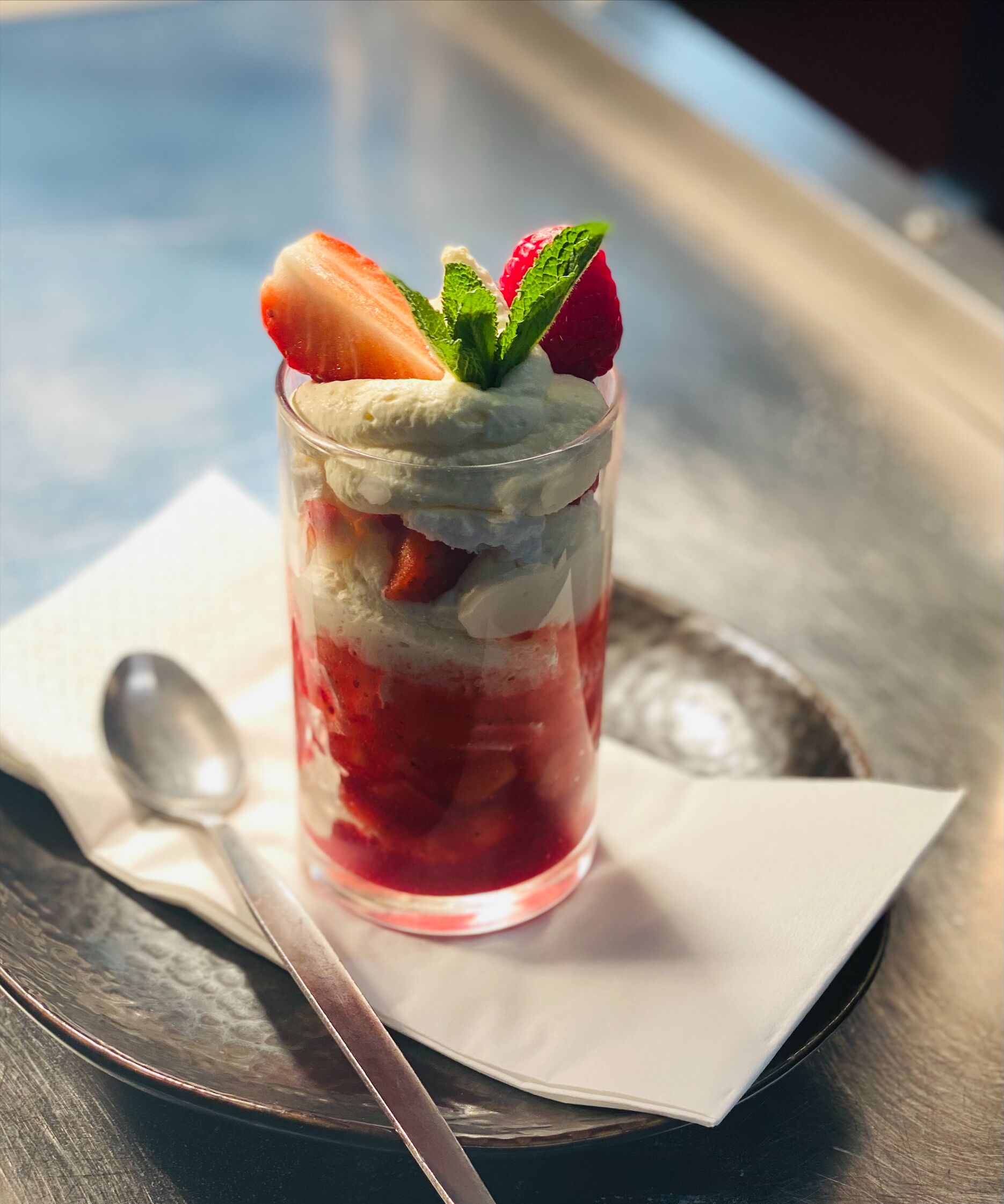 Postre en vaso transparente con fresas, rodajas de plátano y crema batida, servido sobre una bandeja metálica con cuchara y servilleta.