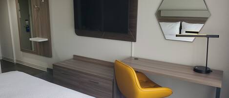 A hotel room with a bed, a desk with a chair, a TV, a mirror, and a wardrobe.