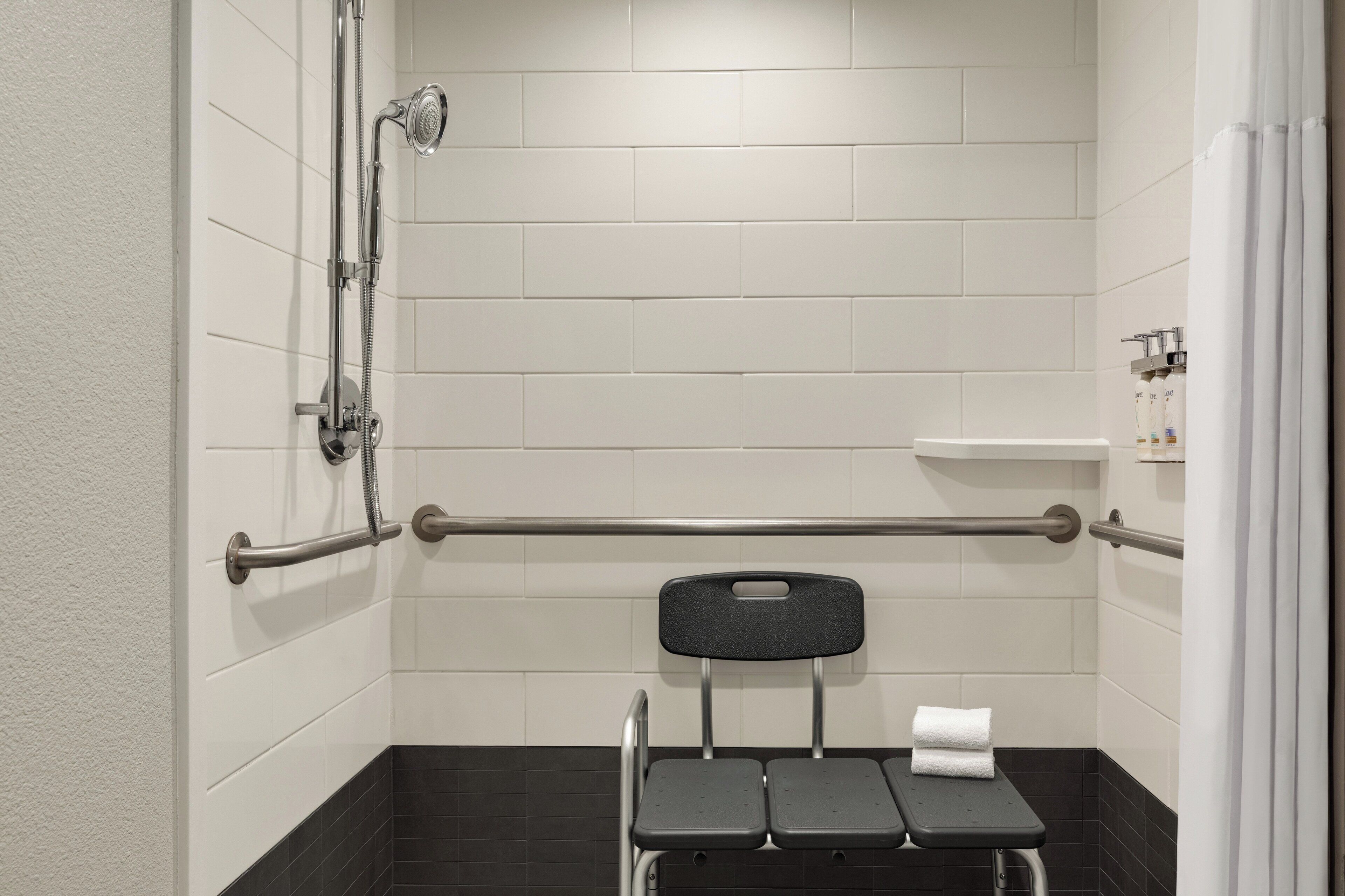 A bathroom with white tiled walls, a black floor, a shower with a handheld showerhead, a grab bar, a black chair with a towel, and a toilet.
