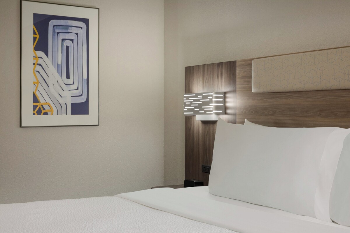 Quarto de hotel com cama bem arrumada, obra de arte na parede e abajur moderno.
