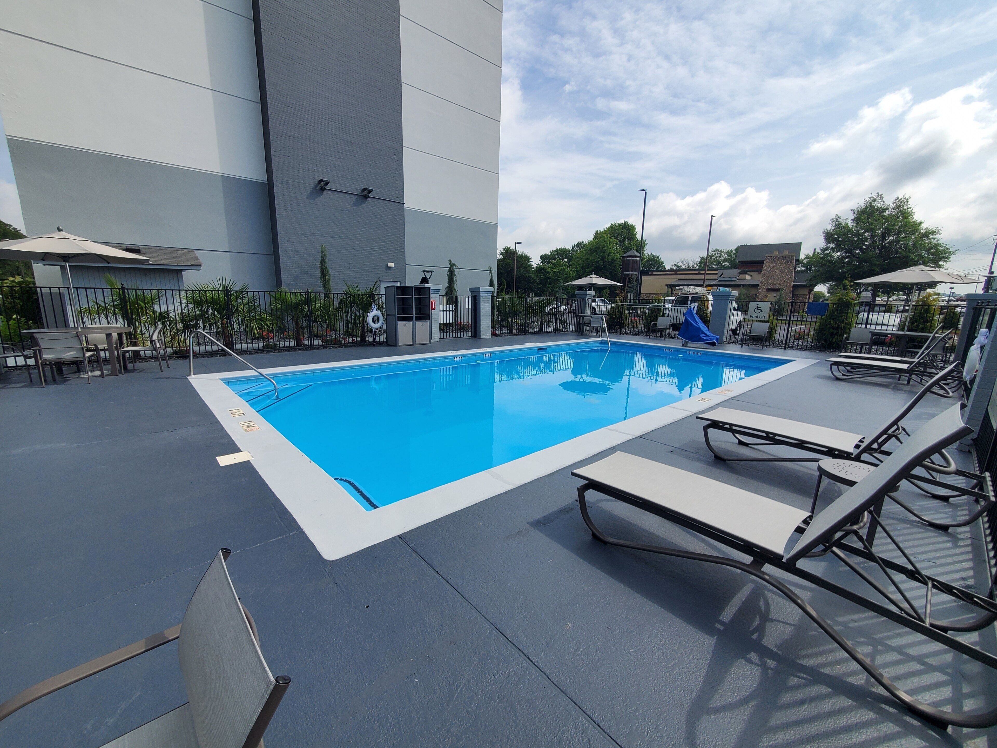 A rectangular outdoor swimming pool with a blue interior, surrounded by lounge chairs and a paved deck.