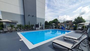 A rectangular outdoor swimming pool with a blue interior, surrounded by lounge chairs and a paved deck.
