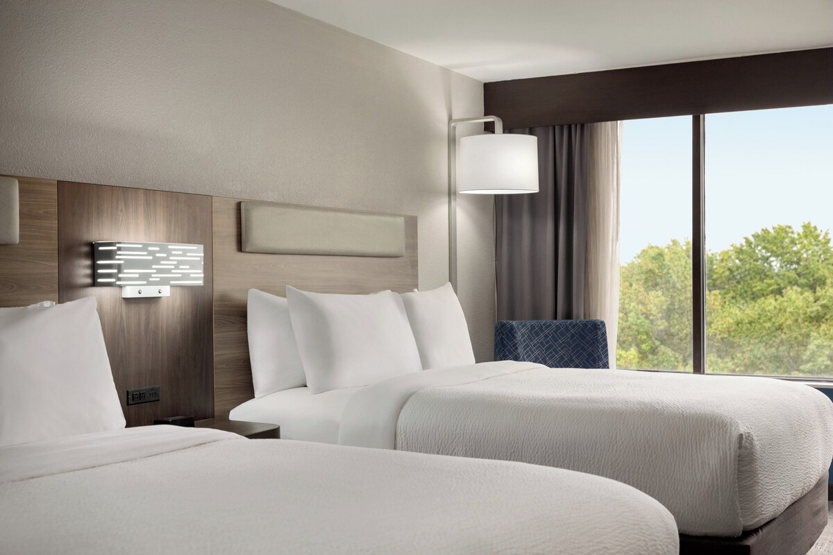 Quarto de hotel com duas camas, luminária moderna fixada na parede e uma grande janela com vista para árvores.