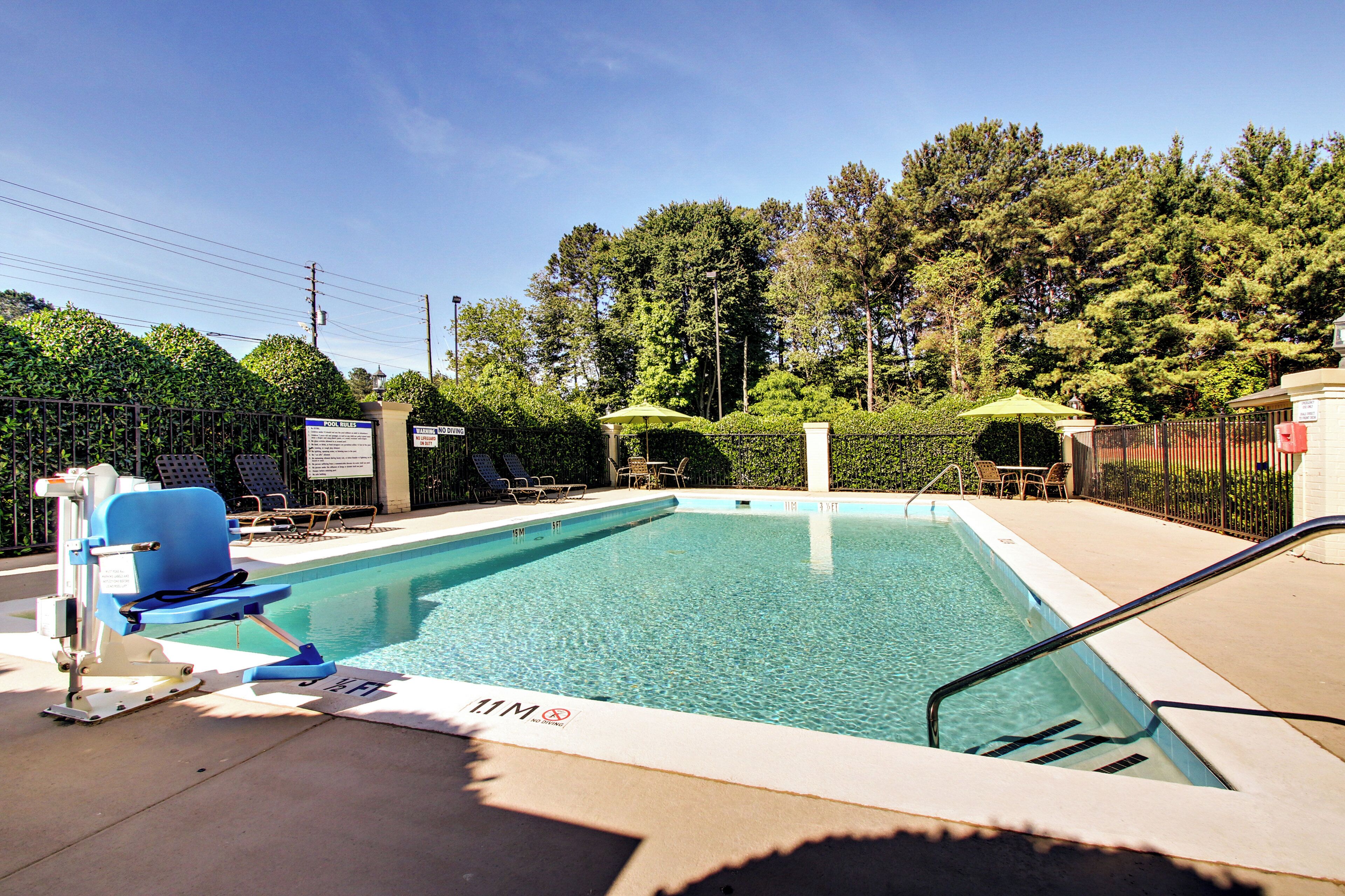 A rectangular pool with clear blue water, surrounded by a metal fence, and equipped with lounge chairs and umbrellas.