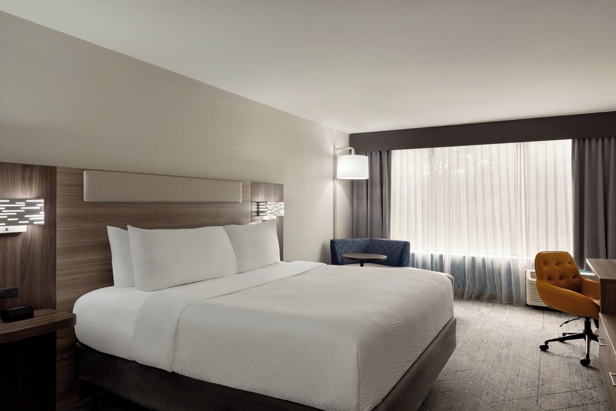 Quarto de hotel com uma cama grande, uma mesa, uma cadeira e uma janela com cortinas.