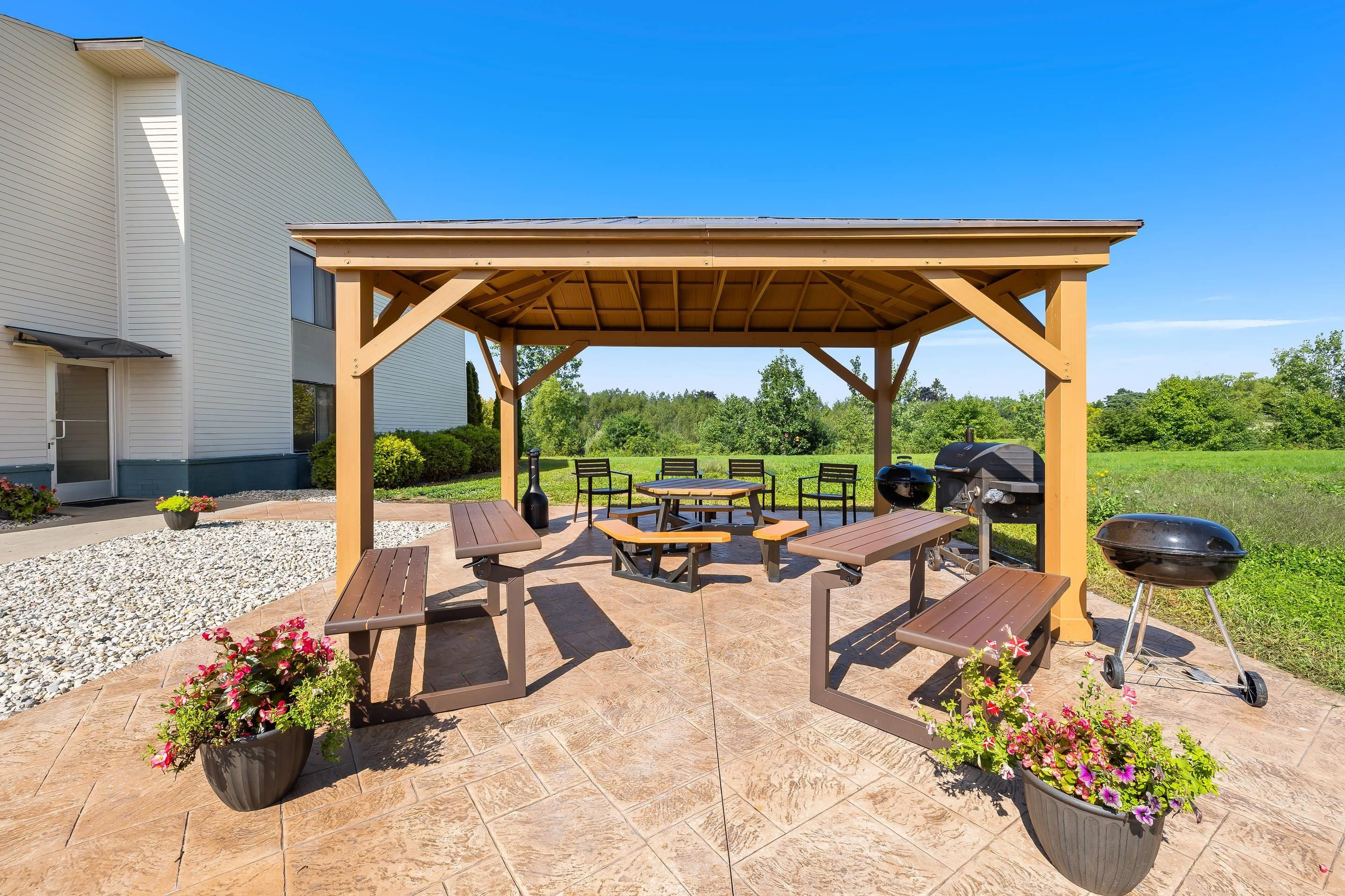 Un patio exterior con una pérgola de madera, mesas de picnic y una zona de barbacoa.