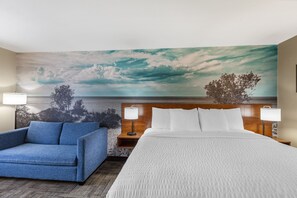 A hotel room with a bed, a blue sofa, a nightstand with a lamp, and a large wall mural of a cloudy sky and trees. - Clarion Pointe Racine - Mount Pleasant (Mount Pleasant)