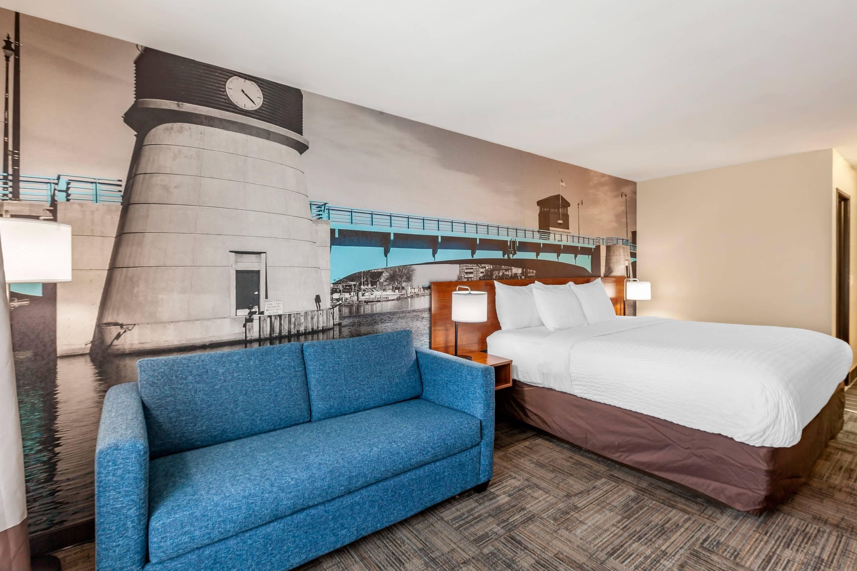 A hotel room with a bed, a blue sofa, a nightstand with a lamp, and a mural of a bridge and tower.