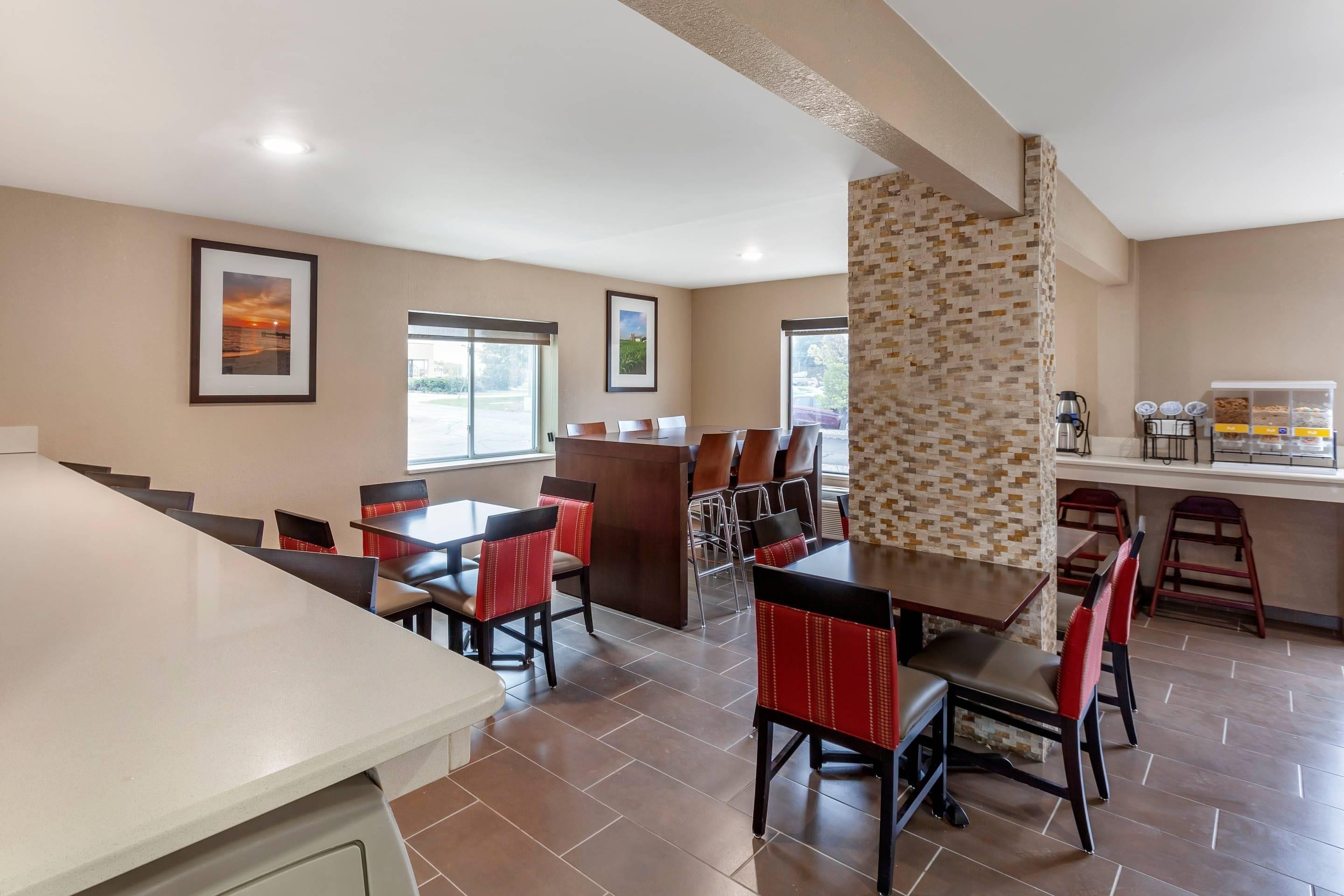 A hotel dining area with multiple tables and chairs, a kitchen area with a coffee station, and framed pictures on the walls.