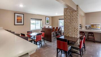 A hotel dining area with multiple tables and chairs, a kitchen area with a coffee station, and framed pictures on the walls.
