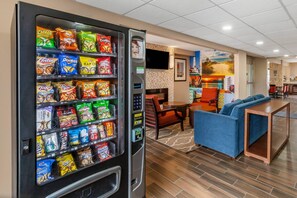 A vending machine stocked with snacks, a seating area with a blue sofa and a small table, and a wall with a beach mural. - Clarion Pointe Racine - Mount Pleasant (Mount Pleasant)