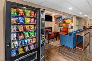 Máquina de vendas com lanches, área de descanso com sofá azul e uma pequena mesa, e parede com mural de praia.