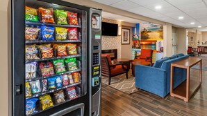 A vending machine stocked with snacks in a hotel lobby with seating and a TV. - Clarion Pointe Racine - Mount Pleasant (Mount Pleasant)