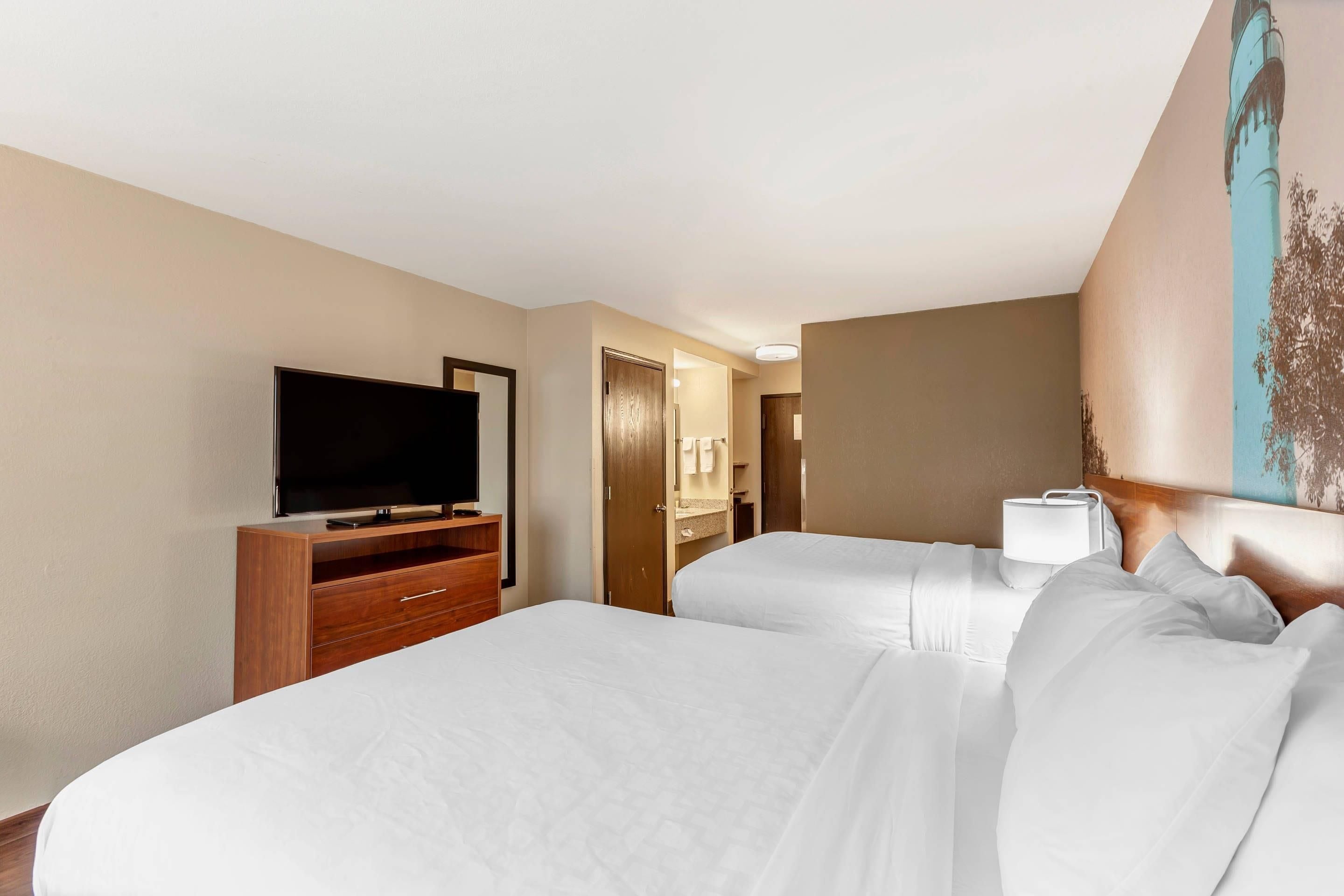 A hotel room with a television on a wooden stand, a bed with white linens, a bedside lamp, and a door leading to a bathroom.