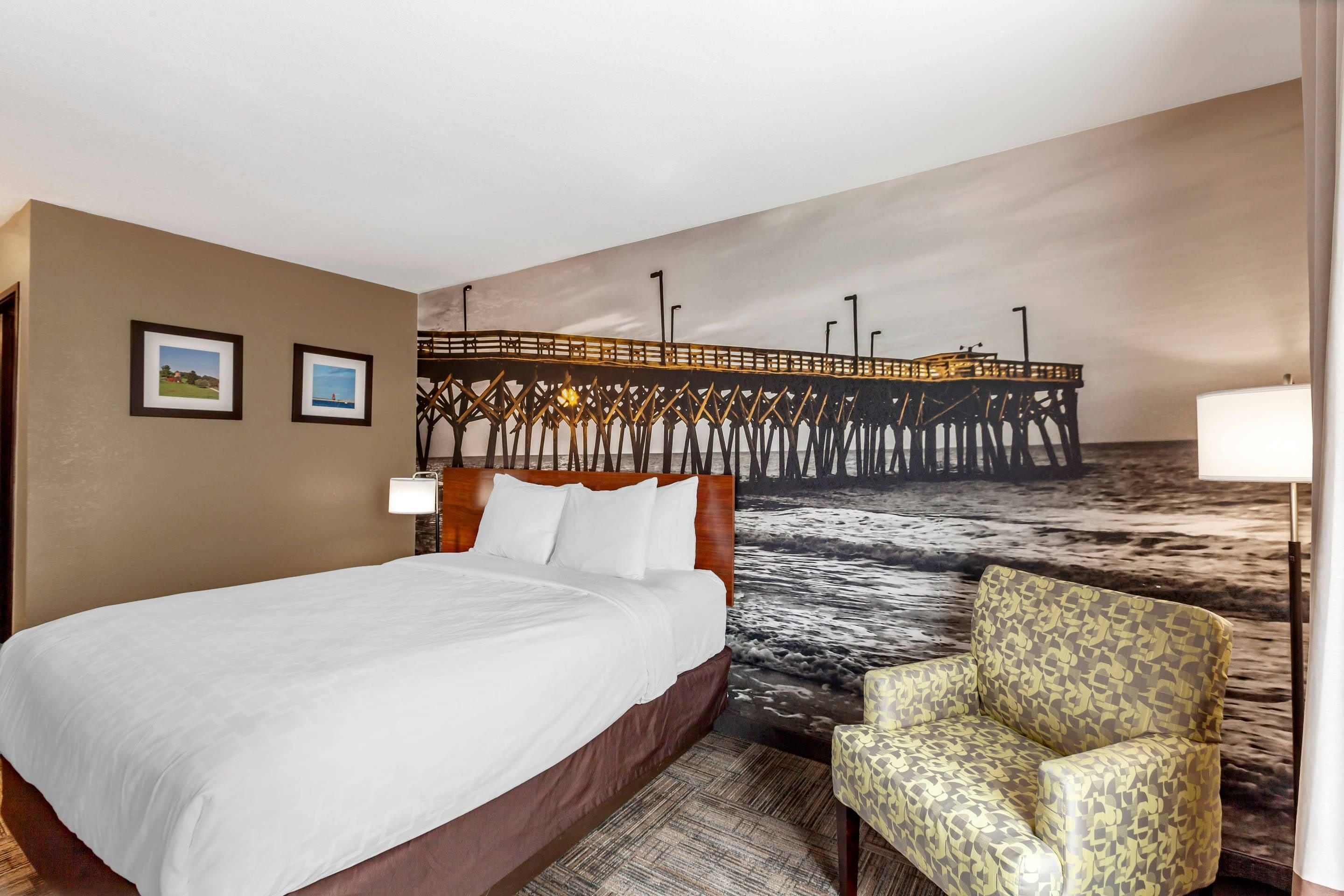 Een hotelkamer met een bed, een stoel, een wanddecoratie van een pier en twee ingelijste foto's.