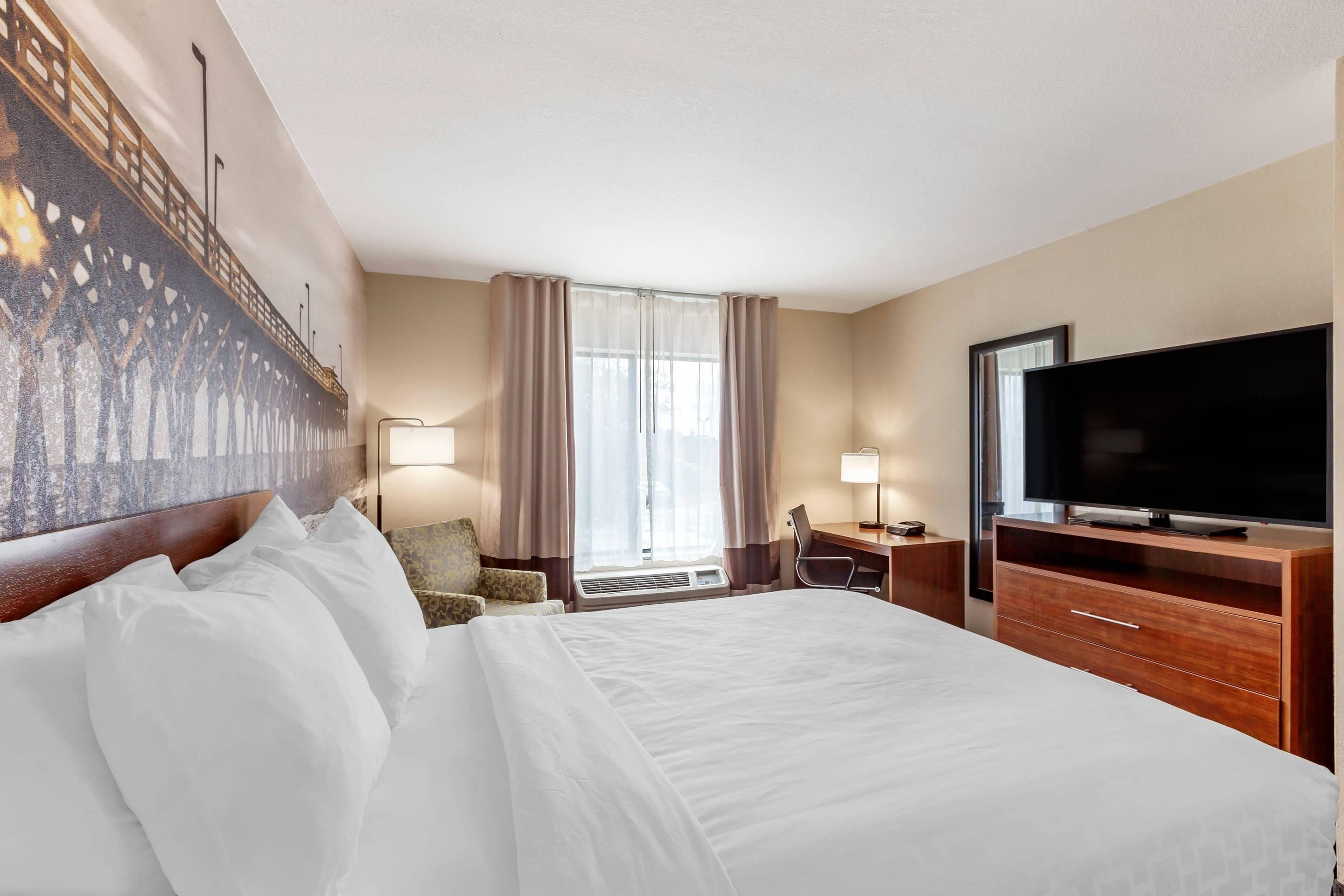 A hotel room with a large bed, a television, a desk with a chair, and a window with curtains.