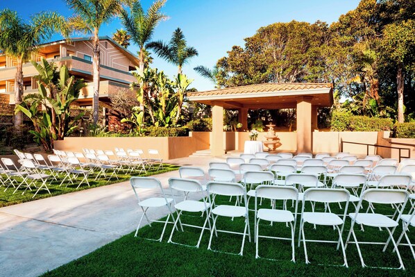 An outdoor wedding setup with white chairs arranged in rows, a gazebo, and a building in the background.