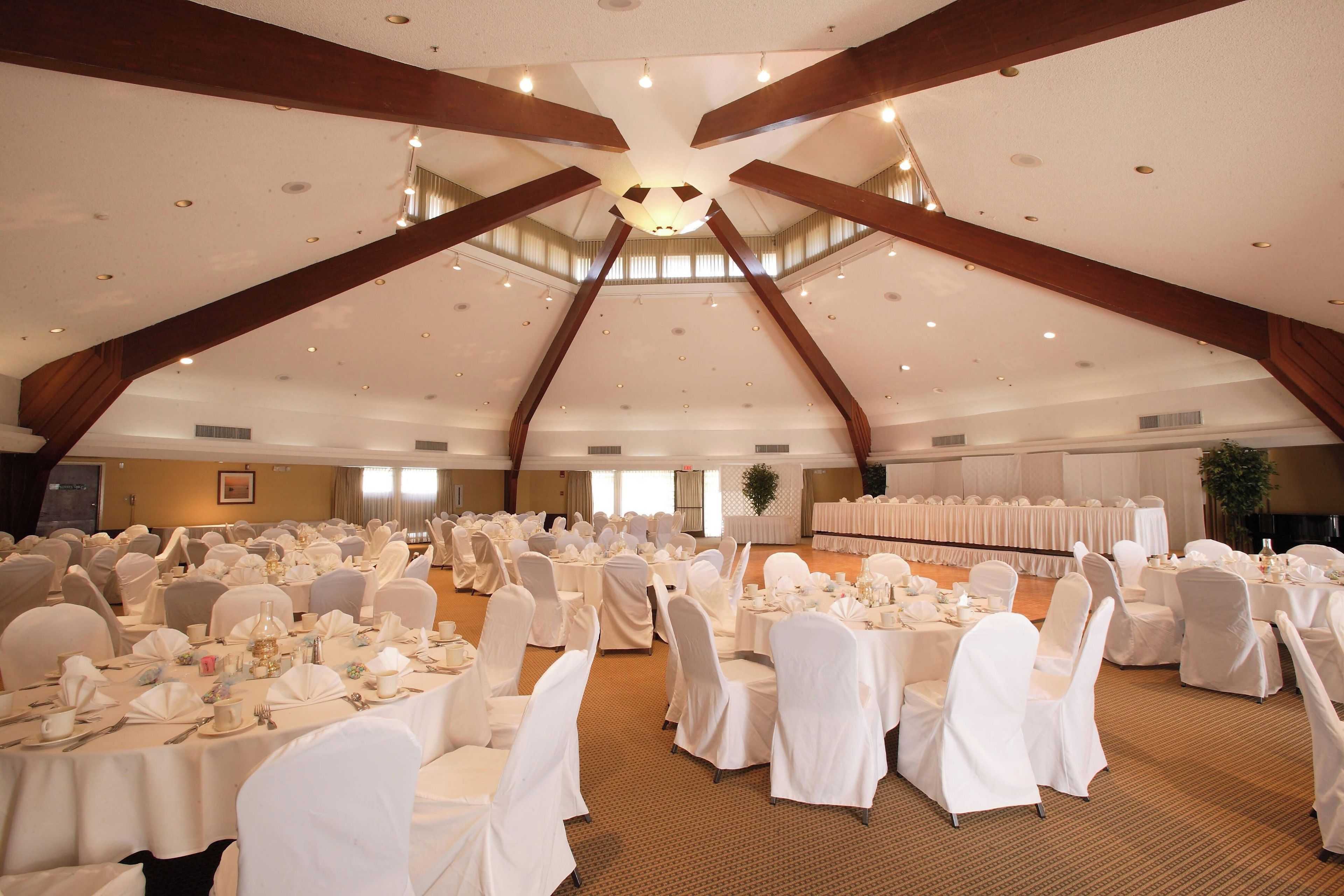 A large banquet hall with high ceilings and wooden beams, arranged with round tables and white-covered chairs.