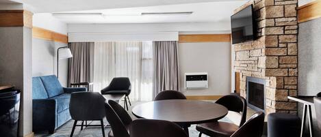 Una habitación de hotel moderna con una mesa redonda de comedor, asientos cómodos, un televisor de pantalla plana montado en una pared de piedra y una chimenea.