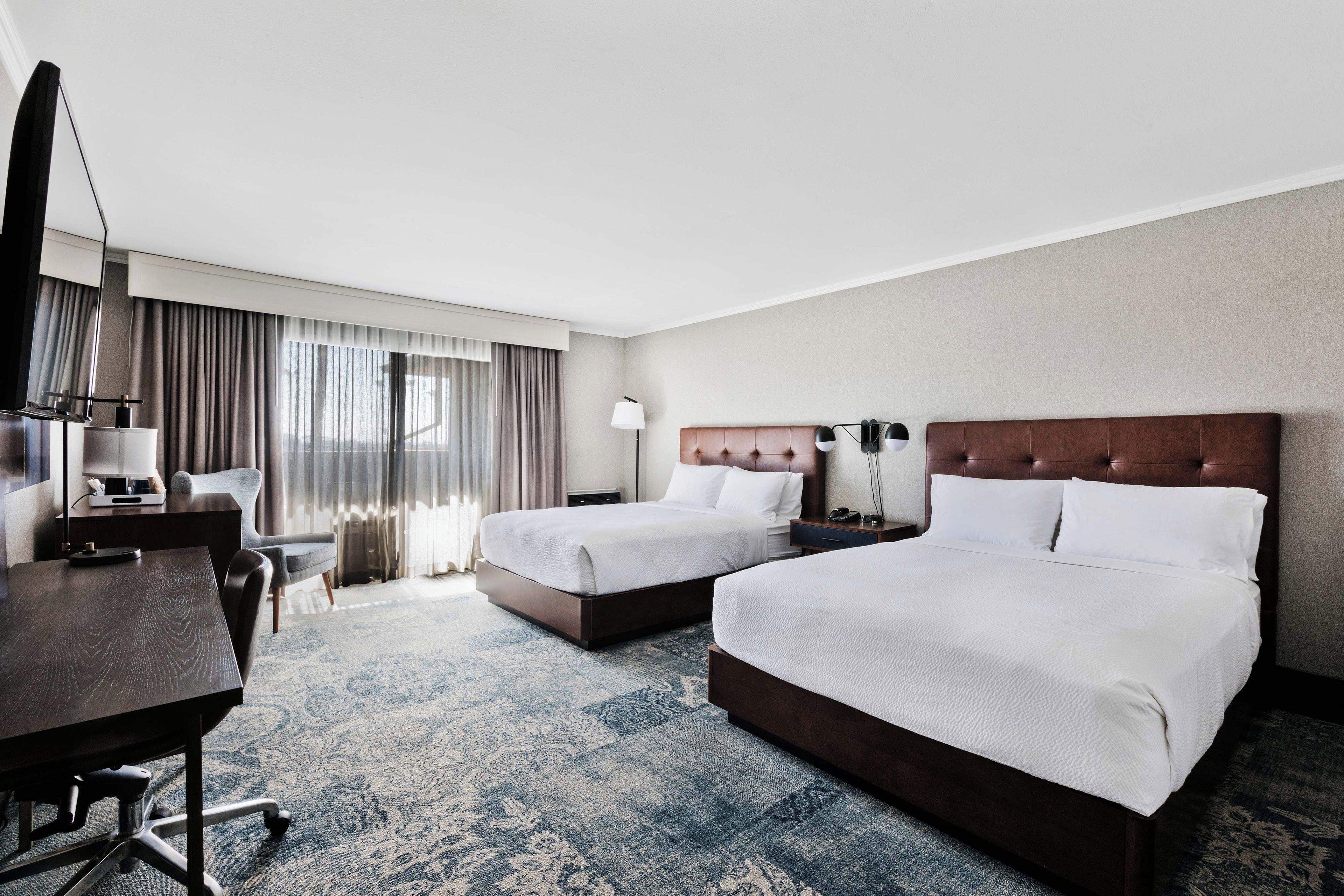 A hotel room with two beds, a desk, a chair, a lamp, and a large window with curtains.