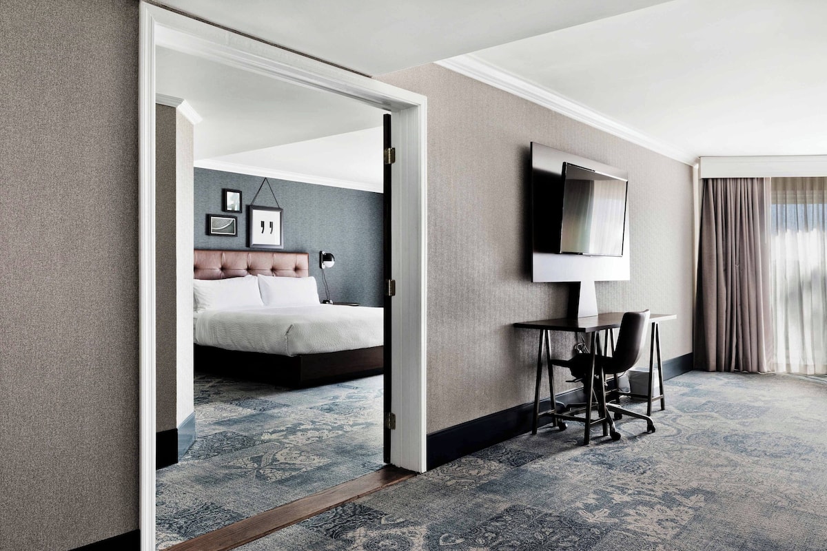 Une chambre d’hôtel avec un lit, un bureau et une chaise, une télévision et une grande fenêtre avec des rideaux.