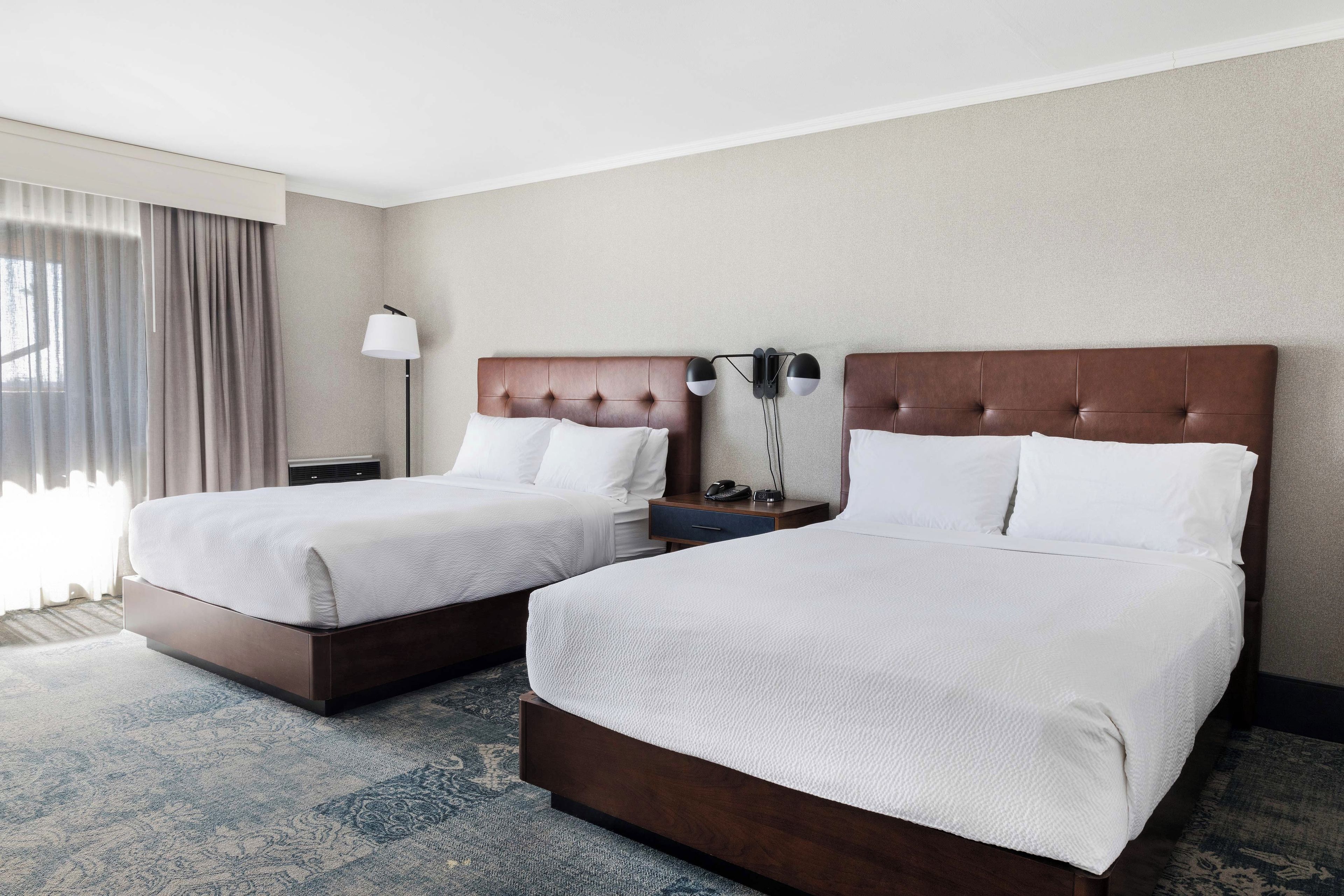 A hotel room with two beds, a nightstand, a lamp, and a view of the outdoors.