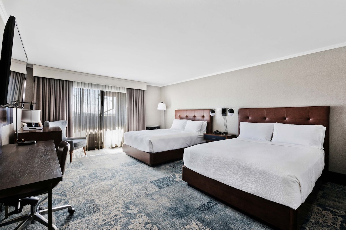 Une chambre d’hôtel avec deux lits, un bureau, une chaise, une télévision et une grande fenêtre avec des rideaux.
