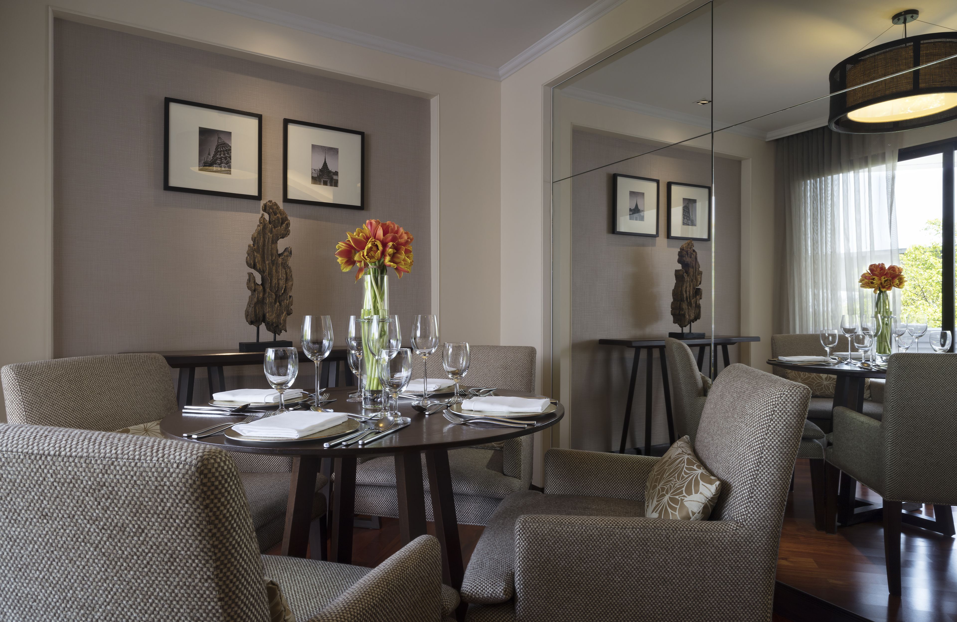 A dining area with a round table set for four, surrounded by upholstered chairs. There are framed pictures on the walls, a vase with flowers on the table, and a large mirror reflecting the room.