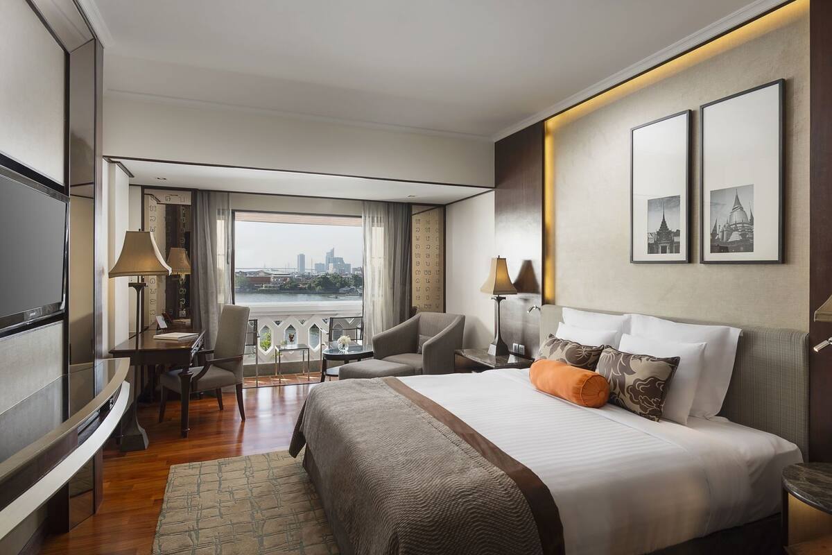 A hotel room with a large bed, a desk with a lamp, a chair, a sofa, and a view of a cityscape through a window.