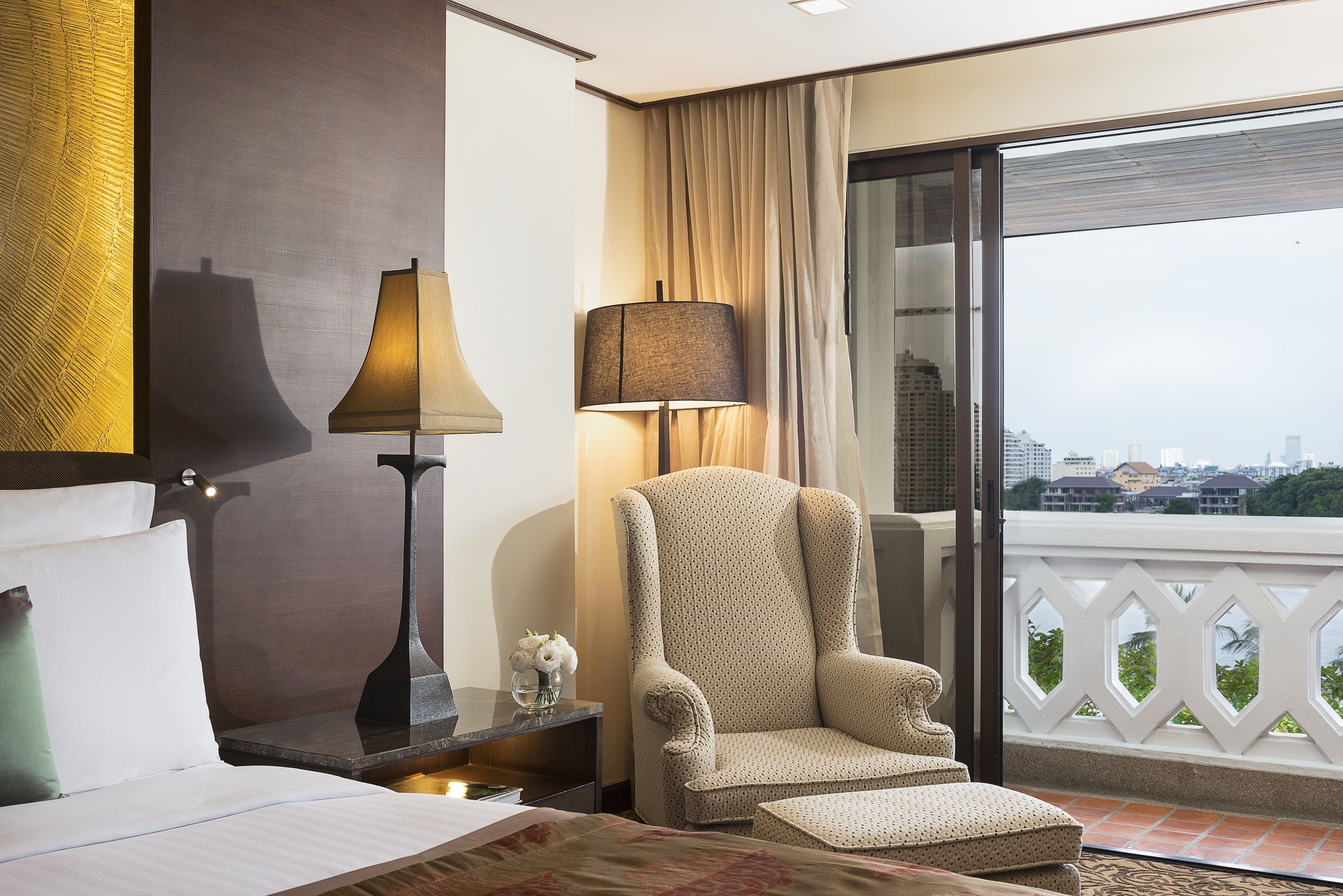 Habitación de hotel con una cama, dos lámparas, una silla y vistas de la ciudad a través de amplios ventanales.