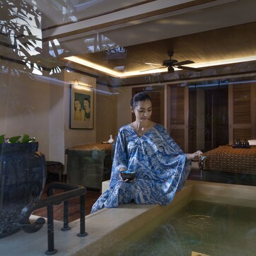 A woman sitting by a bathtub in a well-lit room with a ceiling fan, a bed with a patterned bedspread, and a large window with blinds.