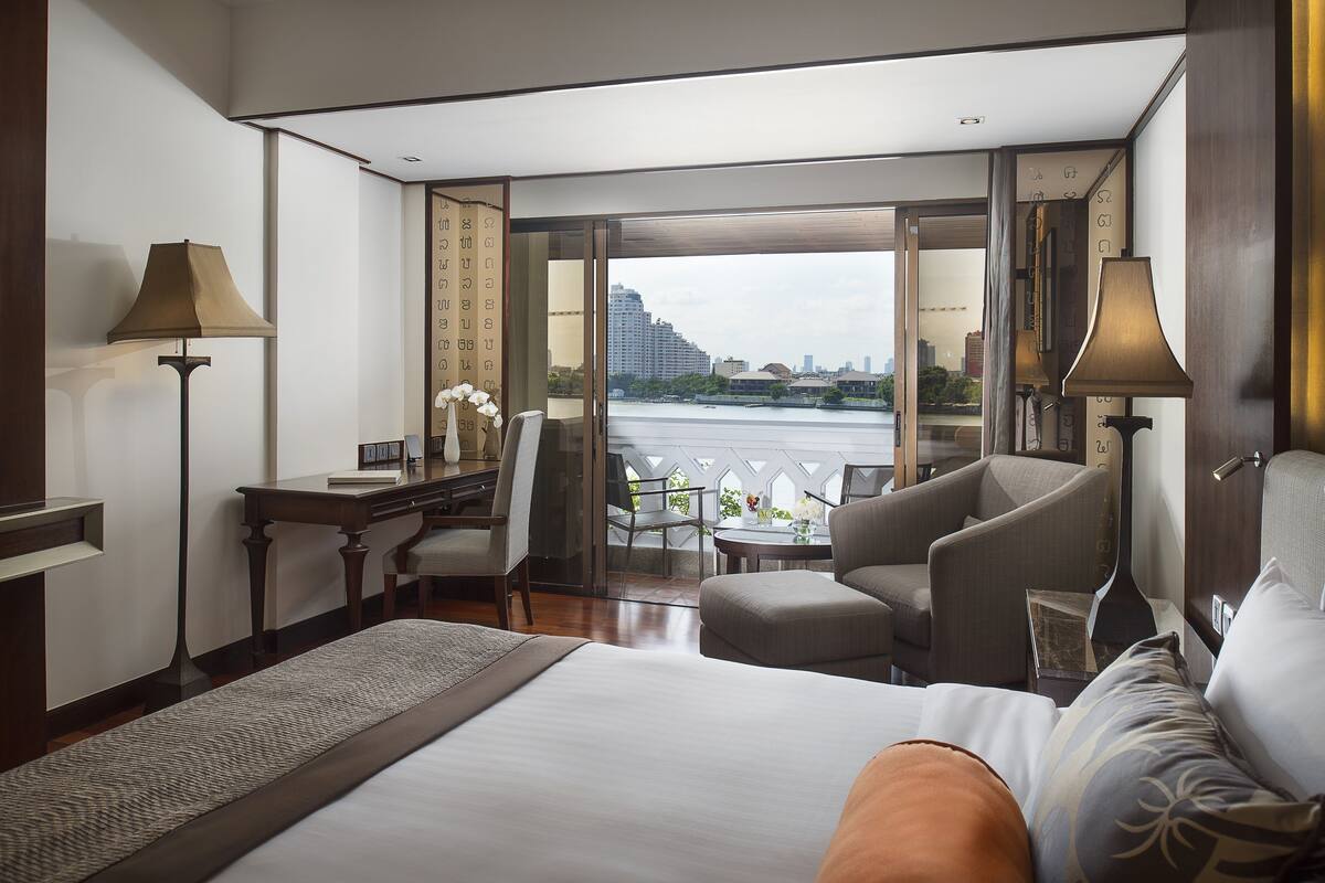 A hotel room with a large bed, a desk, a chair, and a view of a cityscape through a glass door.
